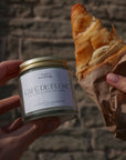 Person holding a jar labeled 'Cafe de Flore' and a croissant on a stone surface.