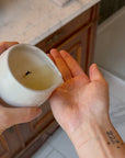Person holding a small ceramic container with a candle inside on a bathroom counter.